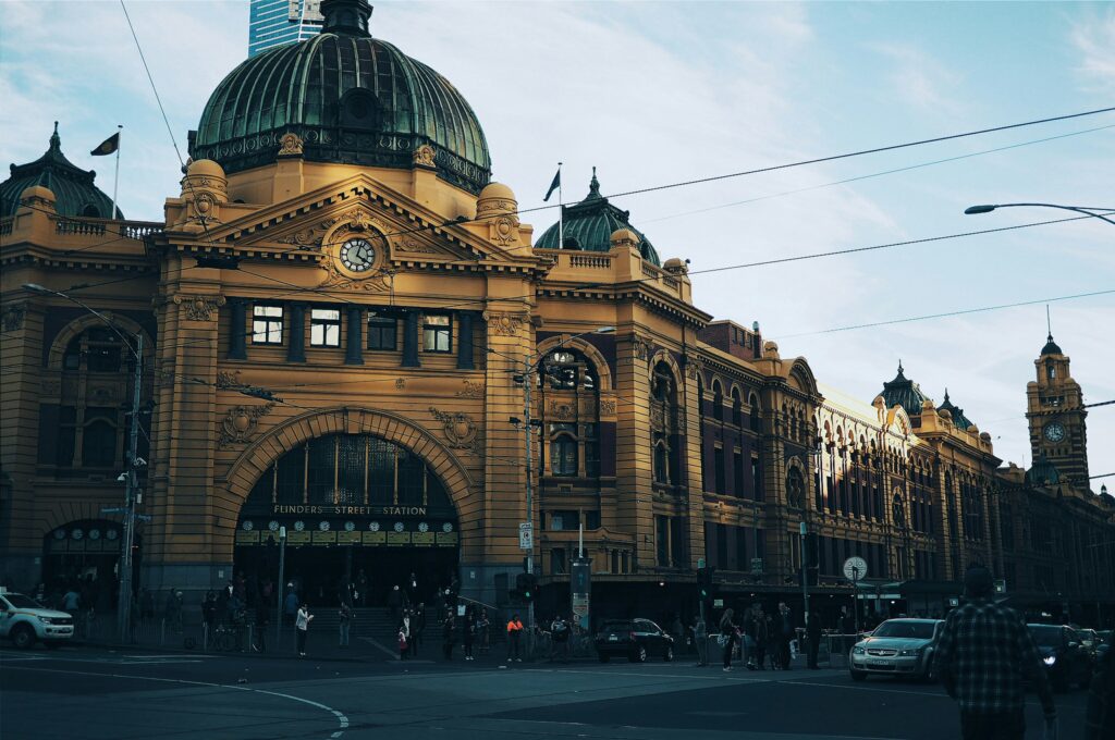 Fotografia da Flinders Street Station em Melbourne, um grande edifício histórico de cor ocre com uma cúpula verde proeminente e vários relógios em sua fachada principal. A rua à frente da estação mostra tráfego de veículos e pedestres. Outros edifícios de arquitetura similar se estendem para a direita, e um céu claro com algumas nuvens é visível acima.