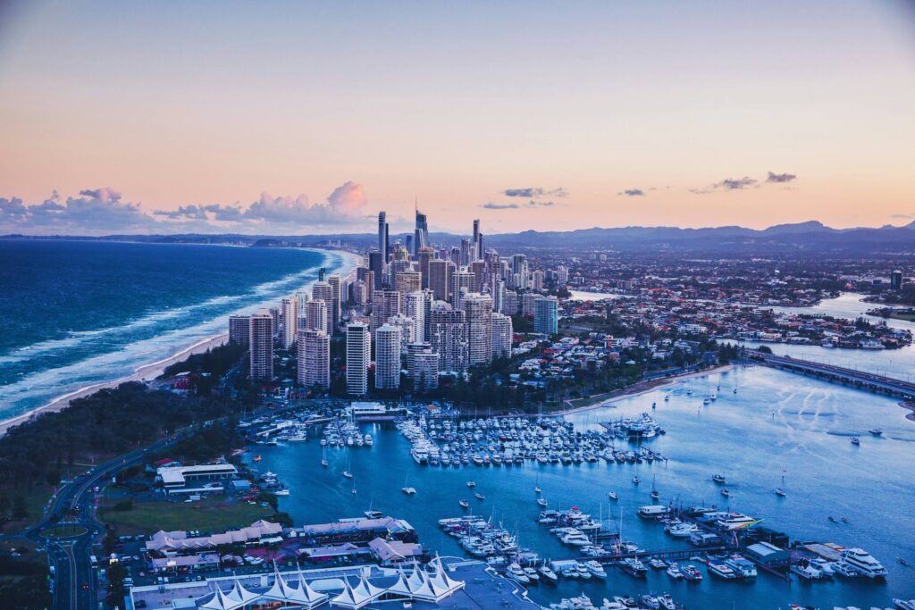Fotografia aérea da Gold Coast, mostrando uma densa área urbana de arranha-céus paralela a uma longa faixa de praia e o Oceano Pacífico. Em primeiro plano, há uma grande marina repleta de barcos e iates, com estruturas baixas em sua margem. No lado direito, um rio ou canal com uma ponte conecta diferentes partes da cidade. O céu ao entardecer exibe tons suaves de azul e rosa, com montanhas distantes no horizonte à direita.