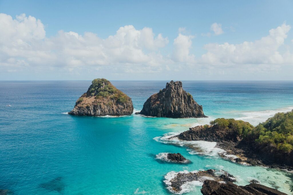 Vista aérea de uma paisagem costeira de Fernando de Noronha, com destaque para a formação rochosa dos Morros Dois Irmãos e as águas cristalinas ao seu redor.