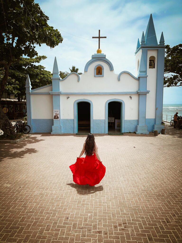 Uma mulher de costas, vestindo um longo vestido vermelho vibrante que esvoaça levemente, caminha em direção à Capela de São Francisco de Assis na Praia do Forte, Bahia. A capela, com sua fachada branca e detalhes em azul claro, possui duas torres pontiagudas e uma cruz no topo. O chão é pavimentado com tijolos em padrão espinha de peixe. Árvores com folhagem verde emolduram a construção, e ao fundo, à direita, é possível ver o mar e algumas pessoas. Bicicletas estão estacionadas à esquerda da capela. A cena captura a beleza arquitetônica e a atmosfera tranquila do local.