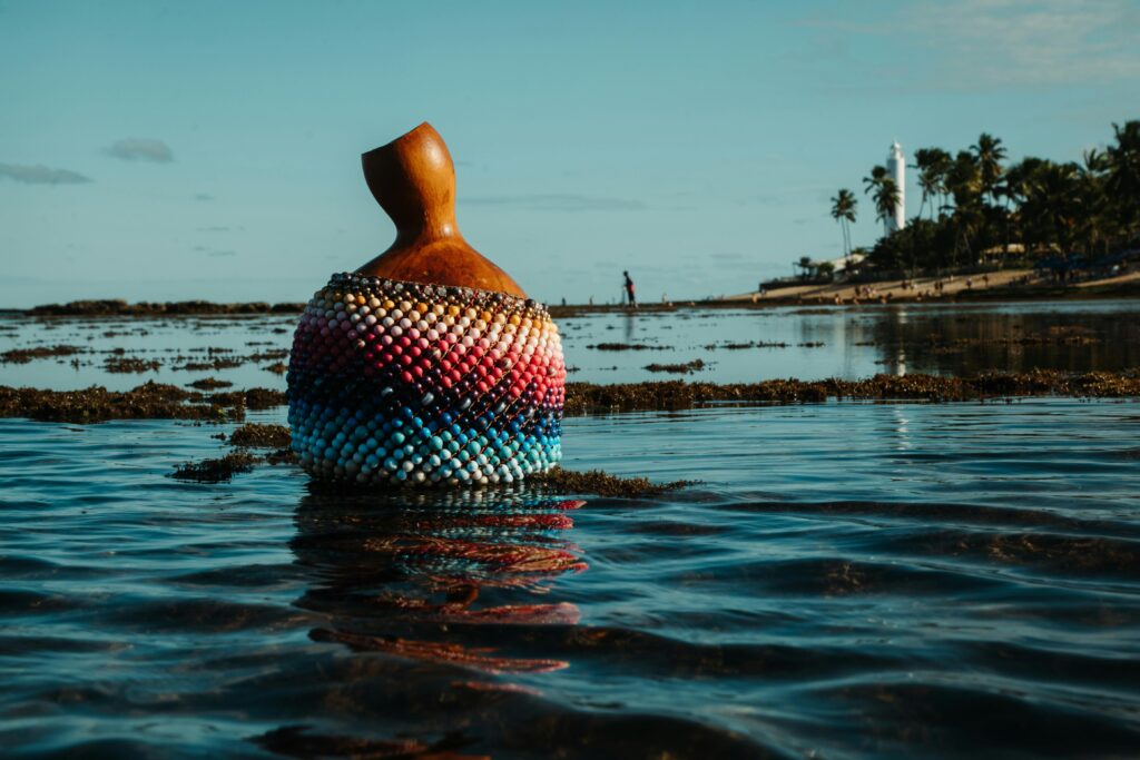 Praia do Forte, Bahia: Um caxixi, instrumento musical de percussão artesanal feito de cabaça e miçangas coloridas em tons de rosa, azul, branco e preto, está parcialmente submerso na água rasa do mar. A superfície da água reflete as cores do instrumento e cria ondulações suaves. Ao fundo, em uma perspectiva de baixa profundidade de campo, observa-se a areia da praia, um coqueiro à direita e o icônico Farol Garcia D'Ávila ao centro, sob um céu azul claro com algumas nuvens brancas. A imagem evoca a cultura afro-brasileira e a serenidade da paisagem litorânea da Praia do Forte.