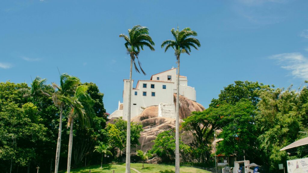 Uma fotografia do Convento da Penha, um edifício branco em estilo colonial, construído no topo de uma colina rochosa e cercado por densa vegetação verde sob um céu azul claro. Duas altas palmeiras em primeiro plano emolduram a vista do convento.