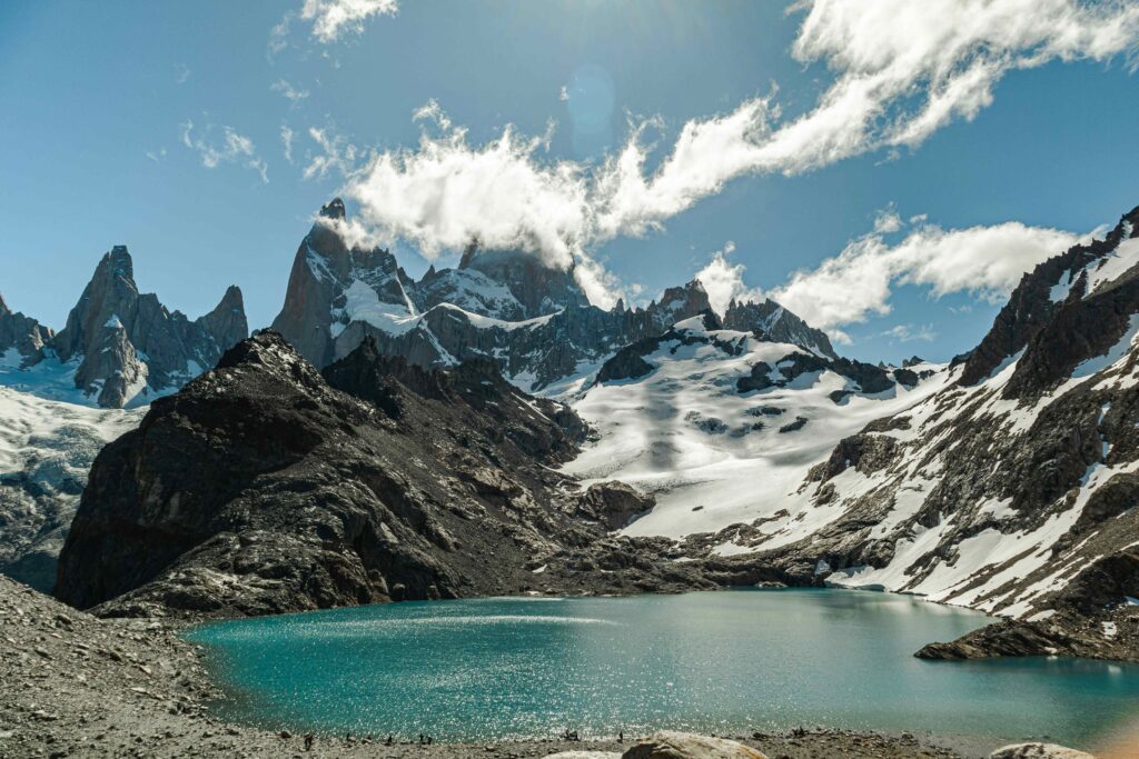 Vista panorâmica da Laguna de los Tres, um lago de água azul-turquesa, cercado por montanhas nevadas e rochosas, incluindo o imponente monte Fitz Roy ao fundo, sob um céu azul com nuvens brancas.