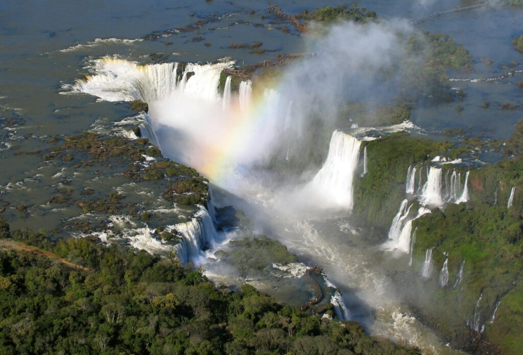 Vista panorâmica das Cataratas do Iguaçu, mostrando a imensidão das quedas d'água e a exuberância da floresta ao redor, sob um céu azul.