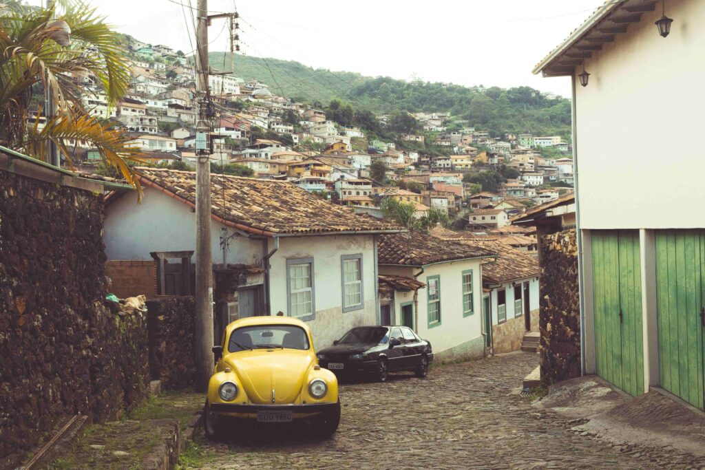 Um fusca amarelo antigo está estacionado em uma ladeira de paralelepípedos em Ouro Preto. Ao lado, há um casarão de arquitetura colonial com telhado de cerâmica e parede branca. No fundo da imagem, pode-se ver uma colina com diversas casas coloridas, características da cidade mineira.