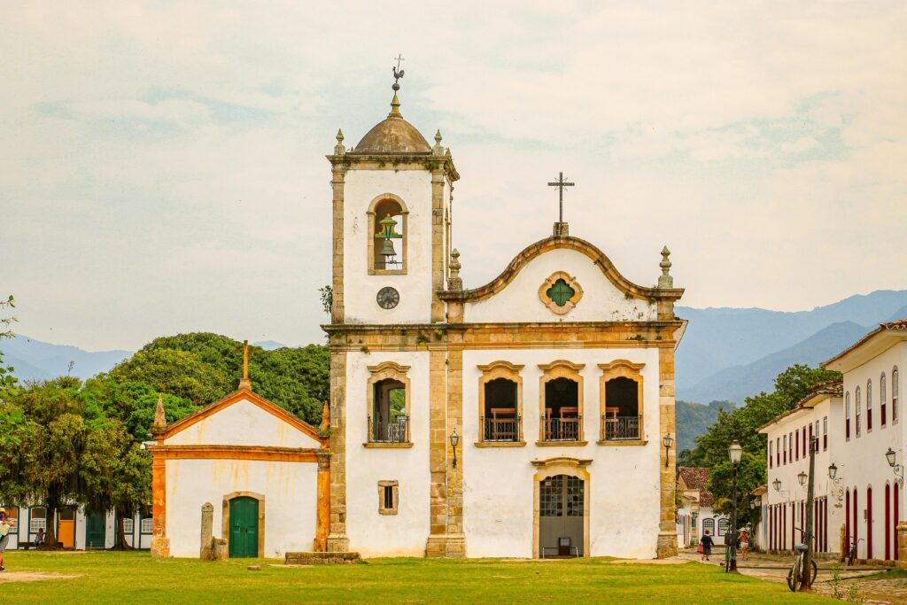Detalhe frontal da Igreja Matriz de Nossa Senhora do Rosário em Paraty, Rio de Janeiro, uma construção colonial branca com campanário e fachada barroca, situada em um gramado verde cercado por edifícios históricos e montanhas ao fundo, ideal para o turismo cultural.