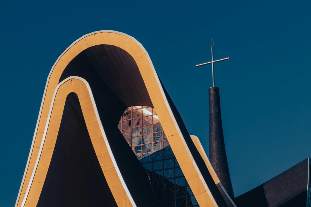 Close-up angular da fachada moderna da Igreja Matriz Nossa Senhora de Guadalupe em Foz do Iguaçu, destacando suas linhas curvas em tom alaranjado, grandes janelas de vidro e uma torre com cruz pontiaguda contra um céu azul intenso.