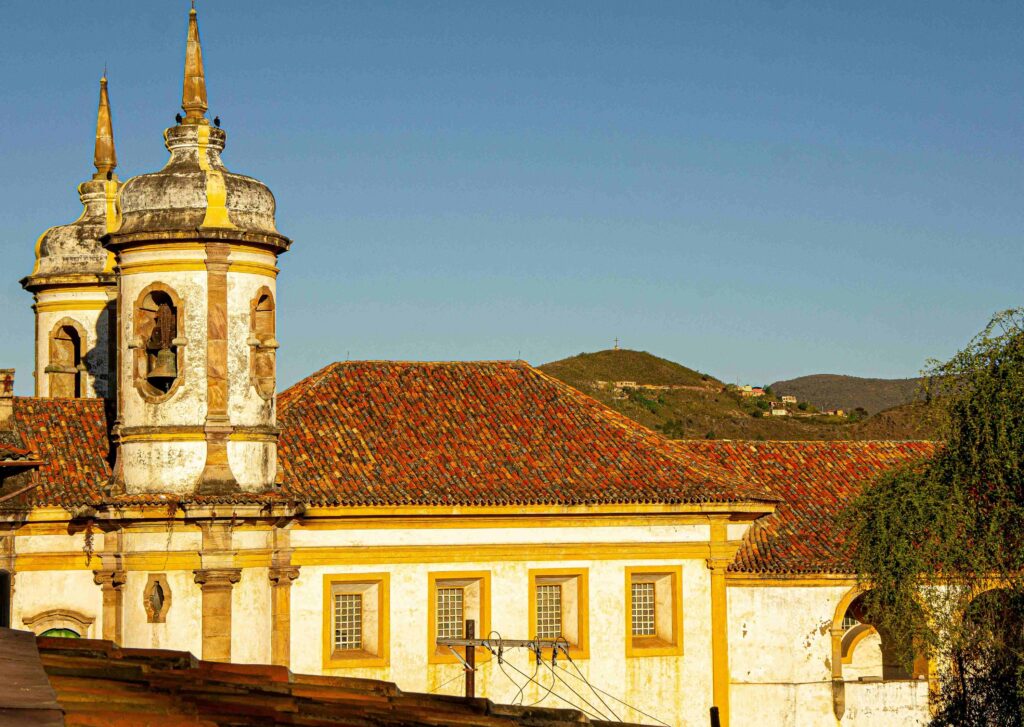 Fachada da Igreja de Nossa Senhora do Carmo, em Ouro Preto, com suas duas torres barrocas em primeiro plano. O edifício colonial, pintado de branco com detalhes em amarelo, tem telhado de cerâmica e está emoldurado por um céu azul. Ao fundo, um morro com vegetação e construções pode ser visto.