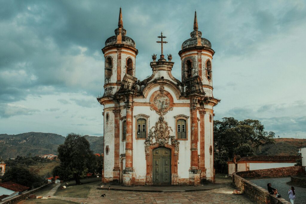 Fachada frontal da Igreja de São Francisco de Assis em Ouro Preto, Minas Gerais, com sua arquitetura barroca em tons de branco e ocre, duas torres sineiras e detalhes ornamentados em pedra sabão. O céu nublado realça a beleza da edificação histórica.