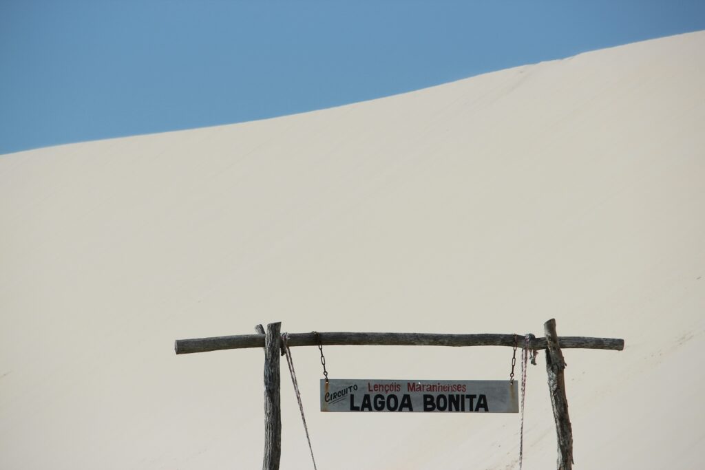 Uma placa de madeira rústica indica o "Circuito Lençóis Maranhenses" e a "Lagoa Bonita", com uma imponente duna de areia clara e céu azul ao fundo, no Maranhão, Brasil.