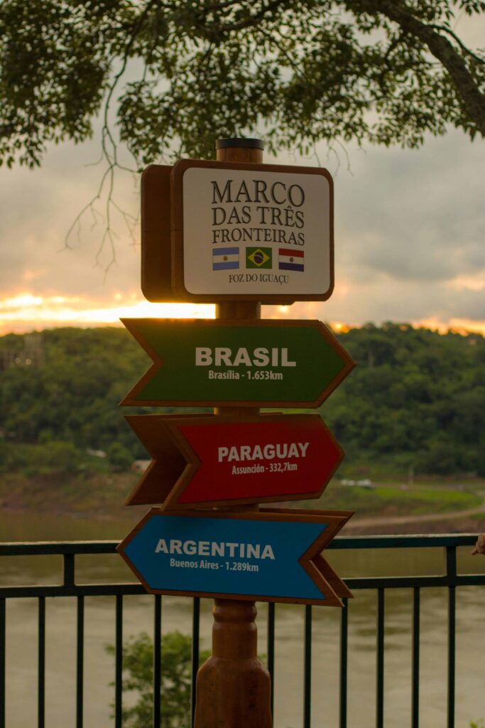 Placa de sinalização de madeira com setas coloridas no Marco das Três Fronteiras em Foz do Iguaçu, apontando para Brasil (Brasília), Paraguai (Assunção) e Argentina (Buenos Aires), com as bandeiras dos três países no topo, sob um céu alaranjado de pôr do sol e árvores ao fundo.