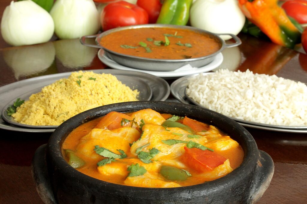 Uma travessa de barro com moqueca capixaba de peixe, colorida e com cheiro verde, servida com arroz branco e farofa, e legumes frescos como tomate, cebola e pimentão ao fundo, em uma mesa de madeira.