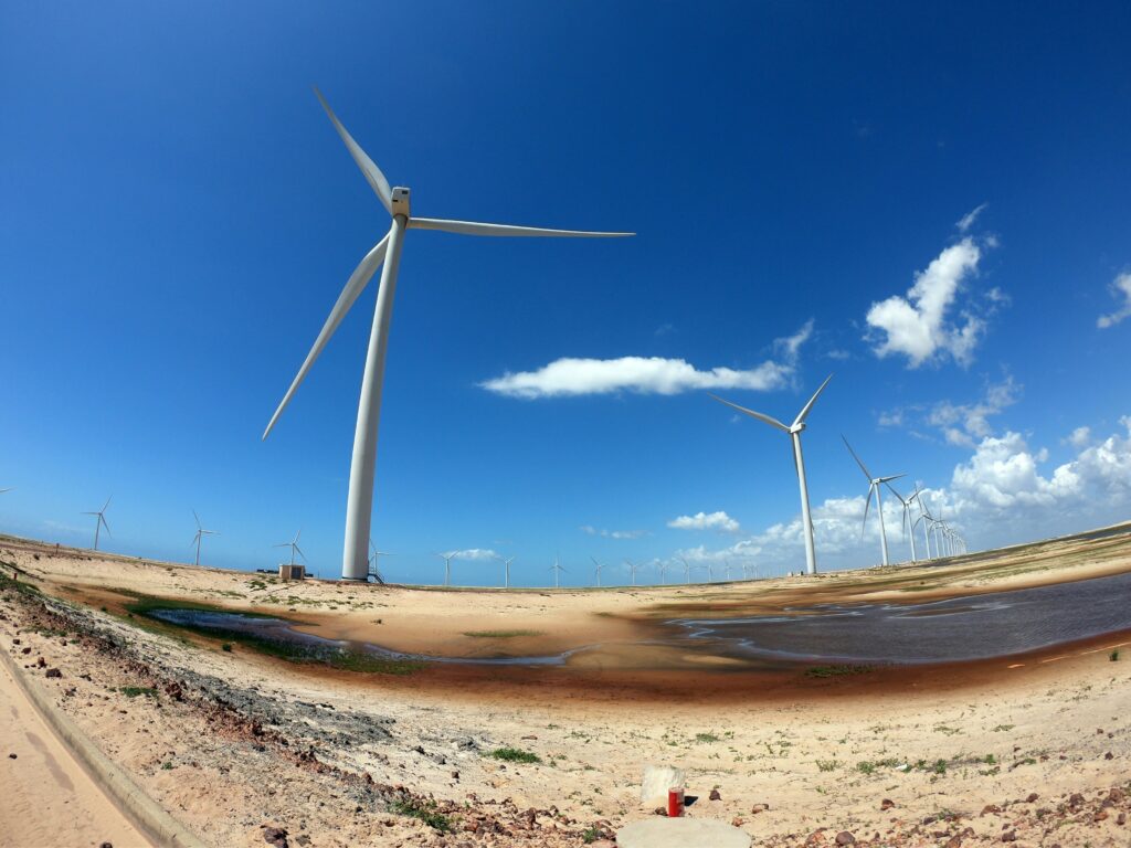Uma fileira de aerogeradores brancos de energia eólica se estende por um terreno arenoso e irregular sob um céu azul vibrante com algumas nuvens brancas, próximo à área dos Lençóis Maranhenses, Brasil.