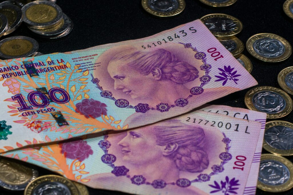 Notas de 100 Pesos Argentinos, ilustradas com a efígie de Eva Perón, dispostas sobre uma superfície escura, com moedas argentinas espalhadas ao redor.