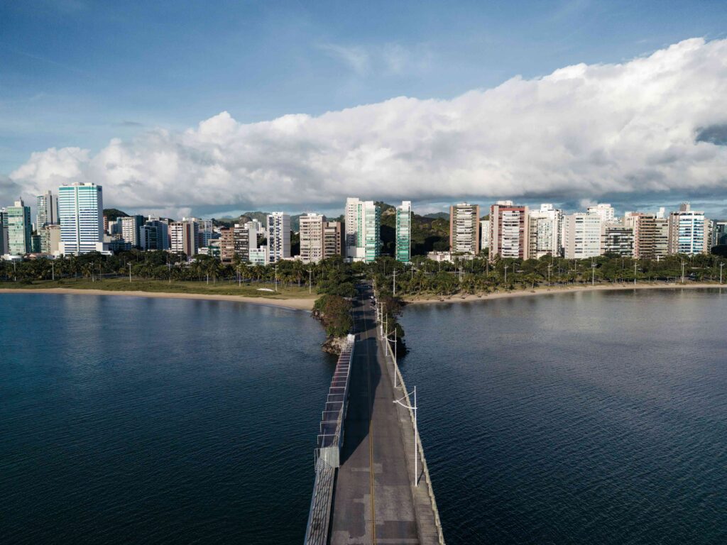 Fotografia aérea da Ponte de Camburi em Vitória, Espírito Santo, atravessando águas calmas em direção a uma extensa faixa de areia com palmeiras e uma densa área urbana de edifícios altos sob um céu parcialmente nublado.