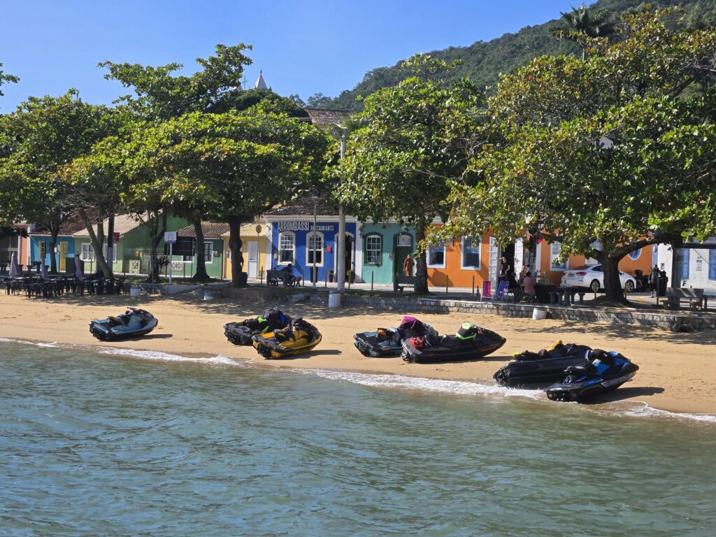 Praia de Paraty com cinco jet skis estacionados na areia clara, em frente a edifícios coloniais coloridos e árvores frondosas, com montanhas verdejantes ao fundo sob um céu azul, destacando o cenário turístico da cidade.