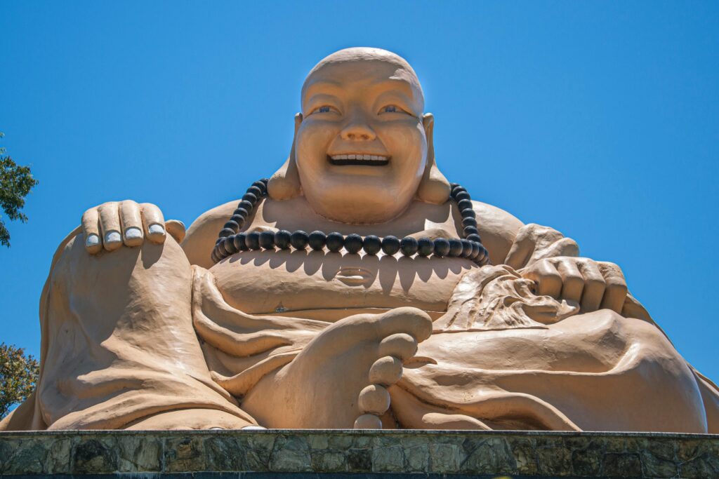 Visão de baixo para cima da gigantesca estátua dourada do Buda Sorridente sentado, com um grande colar de contas escuras, sob um céu azul claro; a imagem foca na expressão alegre do Buda e em sua grandiosidade no Templo Budista Chen Tien em Foz do Iguaçu.