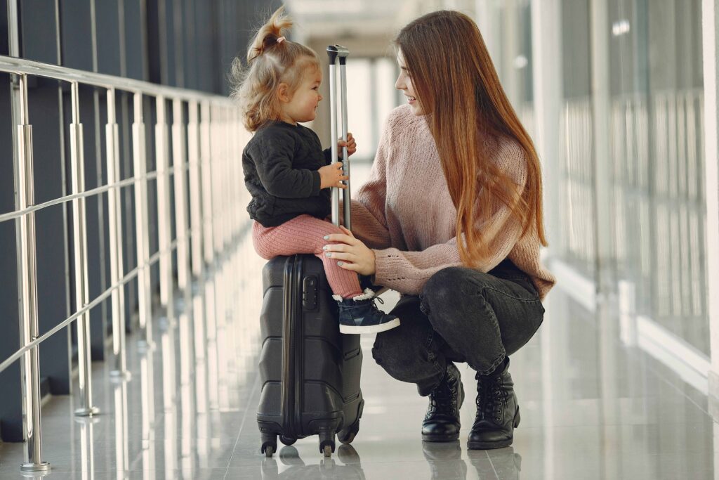 Uma mãe agachada sorri para sua filha pequena, que está sentada em uma mala preta no corredor de um aeroporto, representando a viagem familiar e a importância da autorização para menores.