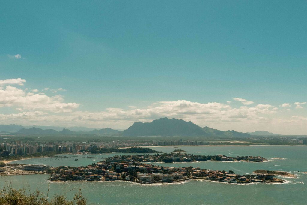Vista panorâmica aérea da cidade de Vitória, Espírito Santo, com ilhas costeiras repletas de edifícios residenciais cercadas por um mar azul-esverdeado, uma extensa área urbana com arranha-céus na orla e uma imponente cadeia de montanhas ao fundo, sob um céu claro com algumas nuvens.