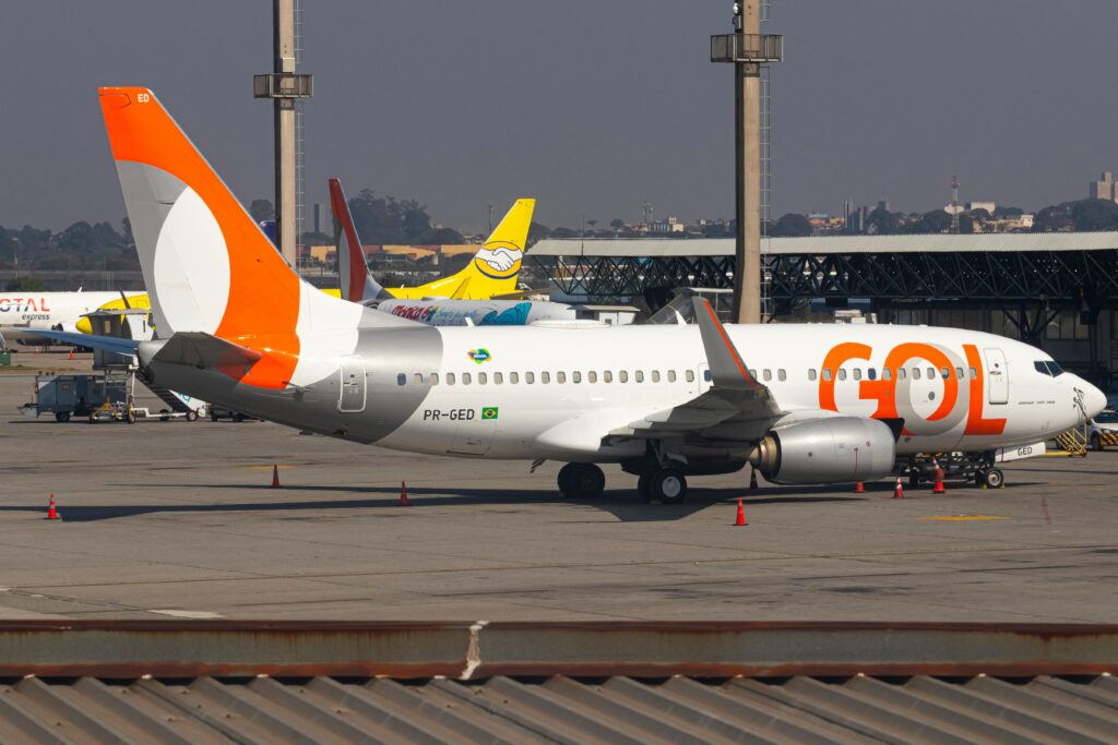 Avião da GOL na pista de um aeroporto brasileiro, com a fuselagem branca e detalhes em laranja e cinza, destacando a asa e a cauda, indicando transporte aéreo e a necessidade de documentação para menores.
