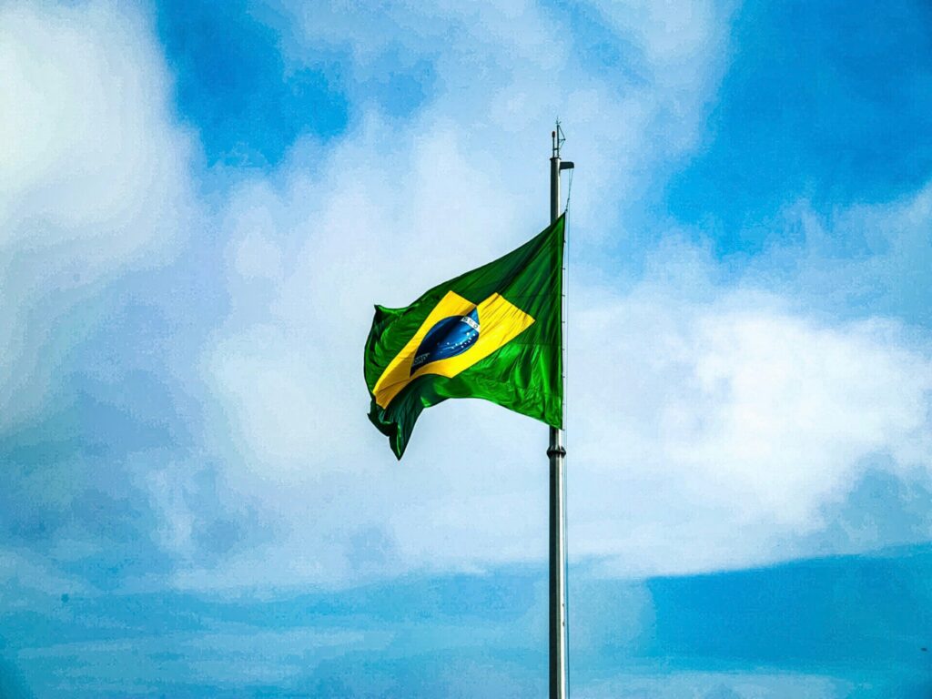 Bandeira do Brasil tremulando em um mastro alto contra um céu azul com nuvens brancas, simbolizando as viagens nacionais e a legislação brasileira sobre autorização de viagem para menores.