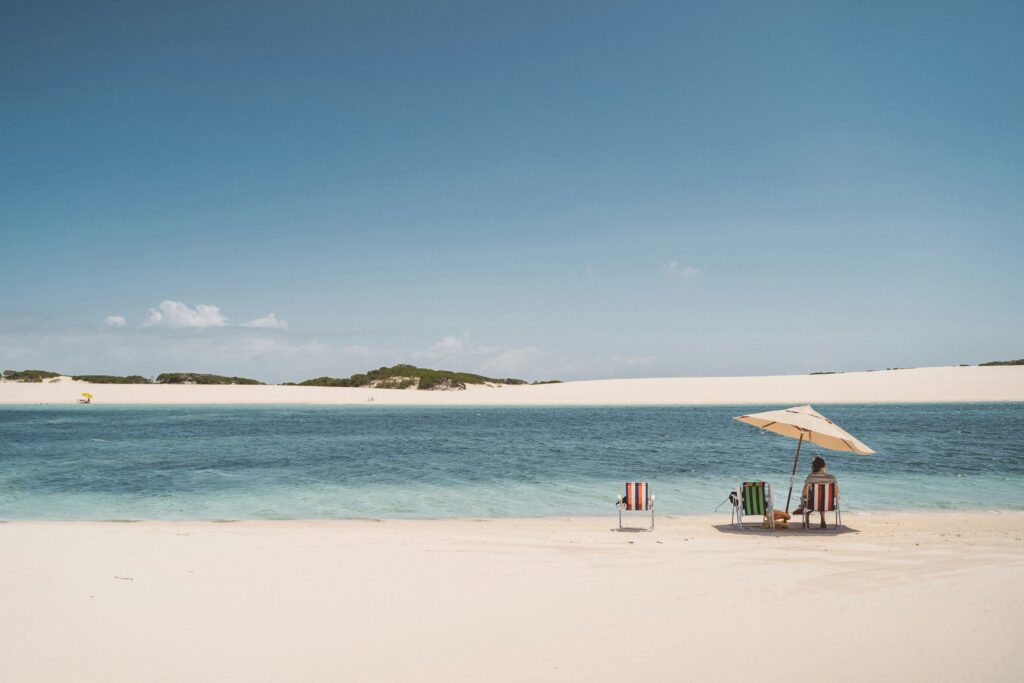 Duas cadeiras de praia listradas e uma pessoa sentada em outra cadeira sob um guarda-sol bege em uma praia de areia clara, à beira de uma lagoa de água azul suave com ondulações, com dunas de areia branca ao fundo e um céu azul claro com poucas nuvens.