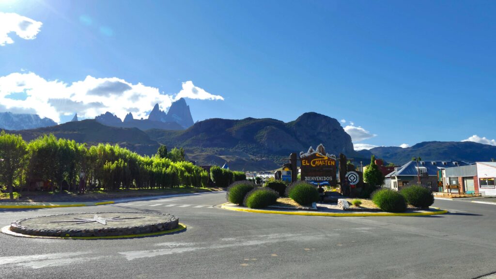 Vista da entrada de El Chaltén com a placa de boas-vindas "El Chaltén, Bienvenidos", um canteiro central com arbustos e o imponente monte Fitz Roy e outras montanhas da Patagônia ao fundo, sob um céu azul e ensolarado.
