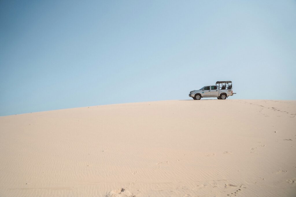 Uma caminhonete 4x4 prata estacionada no topo de uma duna de areia clara sob um céu azul claro nos Lençóis Maranhenses, Brasil.