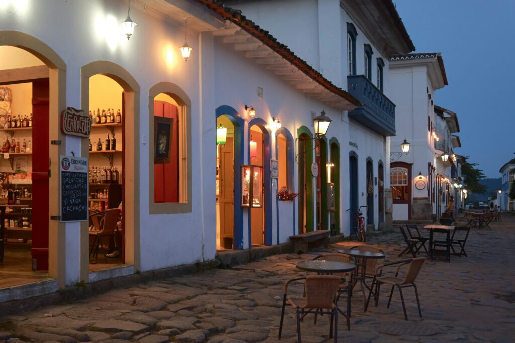 Rua de paralelepípedos no centro histórico de Paraty ao anoitecer, com casarões coloniais brancos e coloridos iluminados por dentro e por luminárias externas, exibindo mesas de bares e restaurantes na calçada, e garrafas em exposição em uma loja à esquerda, destacando a atmosfera noturna e acolhedora da cidade.