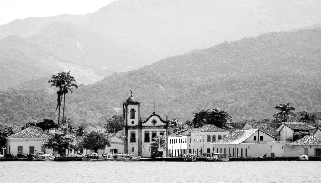 Igreja branca com torre sineira e detalhes em arco, cercada por casas coloniais brancas com telhados de barro, palmeiras esguias à esquerda e barcos ancorados em águas calmas em primeiro plano, com montanhas verde-acinzentadas elevando-se ao fundo sob um céu claro e suave.