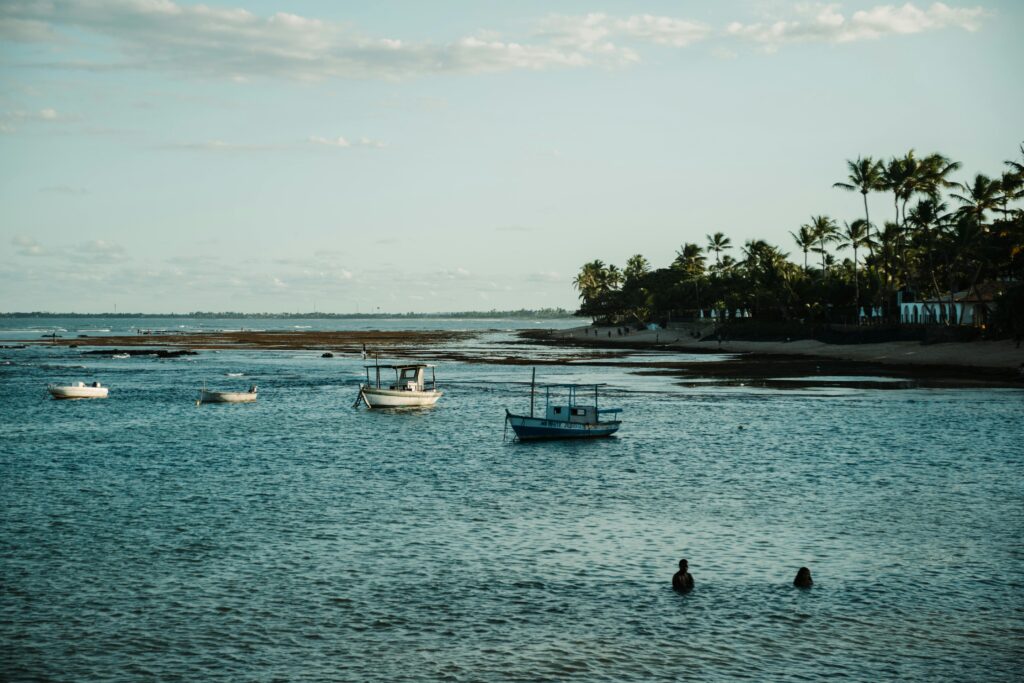 Paisagem costeira da Praia do Forte, Bahia, com suas águas calmas em tons de verde-azulado, onde diversos barcos de pesca estão ancorados. Ao fundo, uma faixa de areia com densos coqueiros delineia a costa, e formações rochosas ou recifes são visíveis na água rasa. No primeiro plano, duas pessoas nadam, desfrutando da serenidade do local sob um céu parcialmente nublado, realçando o ambiente convidativo da praia para lazer e contato com a natureza.