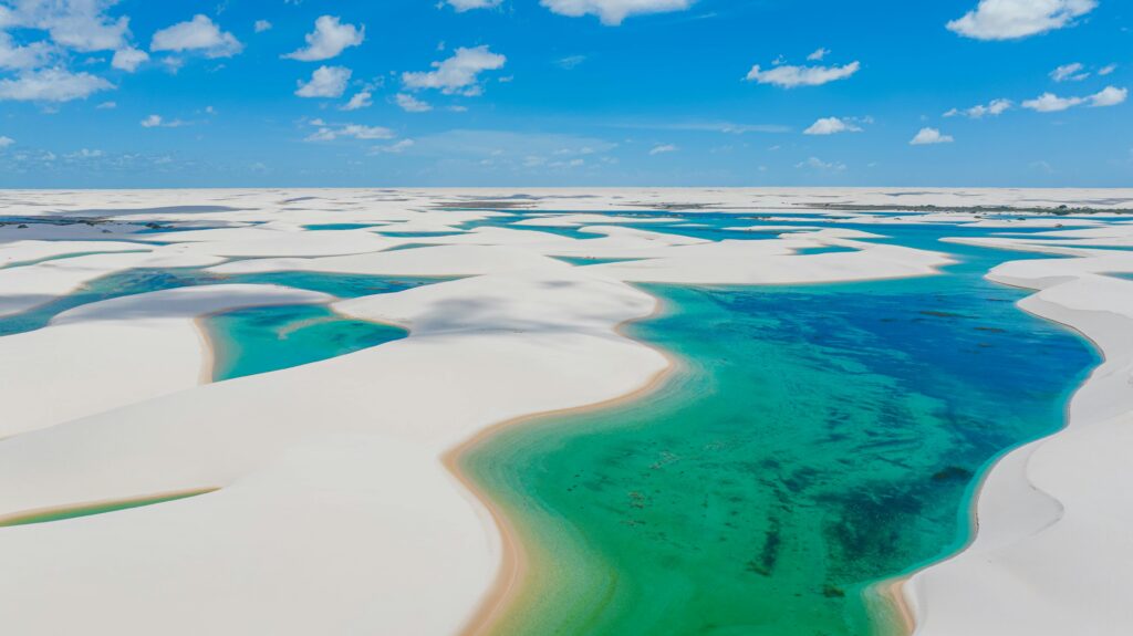 Vista aérea panorâmica dos Lençóis Maranhenses, mostrando inúmeras lagoas de água verde e azul vibrante intercaladas com dunas de areia branca e ondulante sob um céu azul claro com nuvens esparsas.