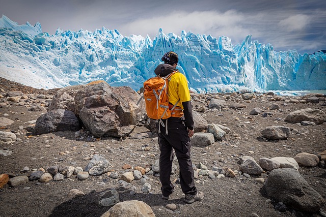 Uma pessoa de costas, usando uma jaqueta amarela e uma mochila de caminhada, observa a vasta parede da geleira Perito Moreno. Ela está em uma trilha de cascalho e pedras grandes, com a imponente parede de gelo azul-claro ao fundo.