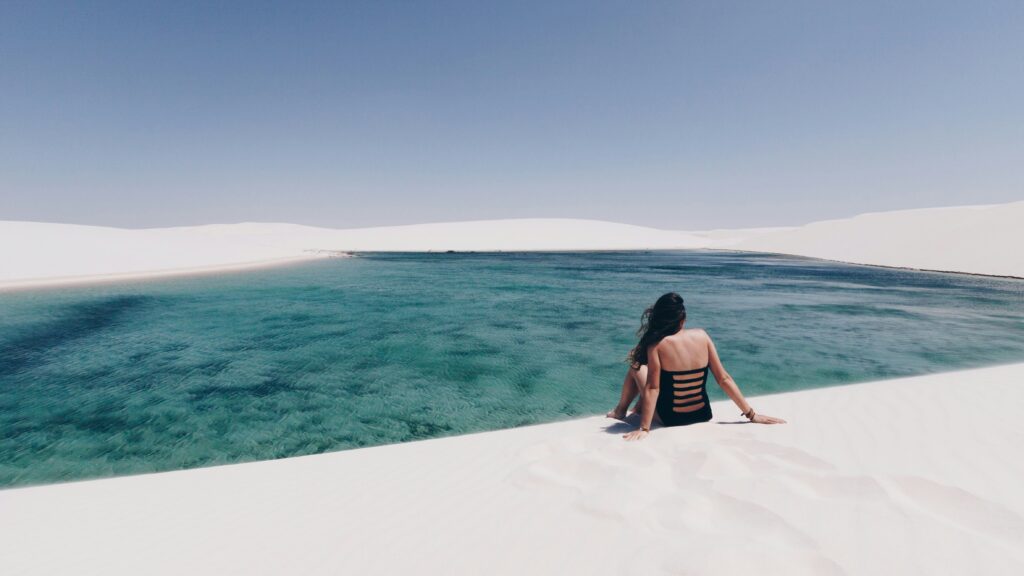 Uma mulher de costas, vestindo um traje de banho preto, está sentada na areia branca de uma duna, observando uma lagoa de água azul-turquesa cercada por outras dunas sob um céu azul claro nos Lençóis Maranhenses, Brasil.