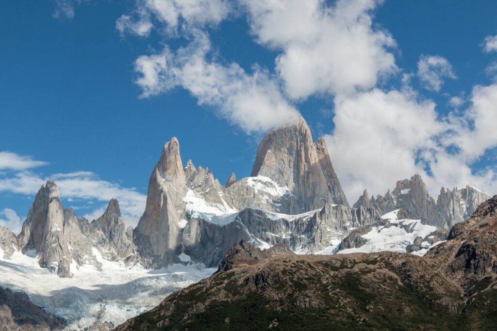 Vista de perto dos picos nevados do monte Fitz Roy e seus arredores em El Chaltén, Argentina, sob um céu azul vibrante com nuvens brancas esparsas. As montanhas rochosas e escarpadas se destacam com gelo e neve em seus vales e encostas.