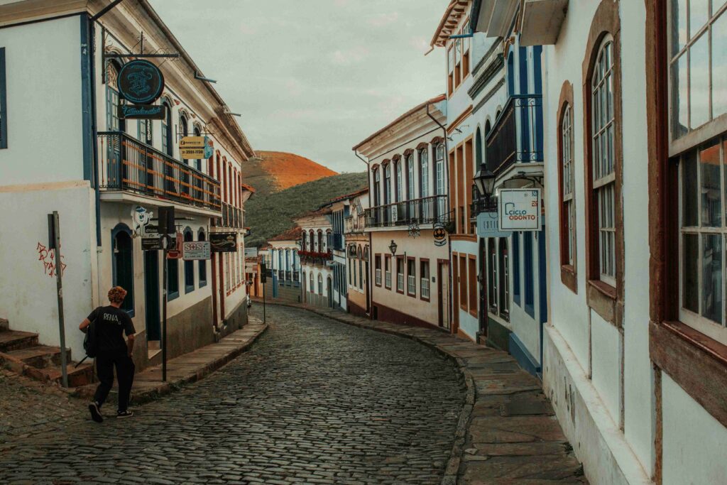 Um homem de costas, vestindo uma camiseta preta, caminha por uma rua de paralelepípedos em Ouro Preto, cercado por casarões coloniais brancos e coloridos com varandas e janelas de madeira. Ao fundo, uma colina coberta de vegetação complementa a paisagem histórica da cidade mineira.