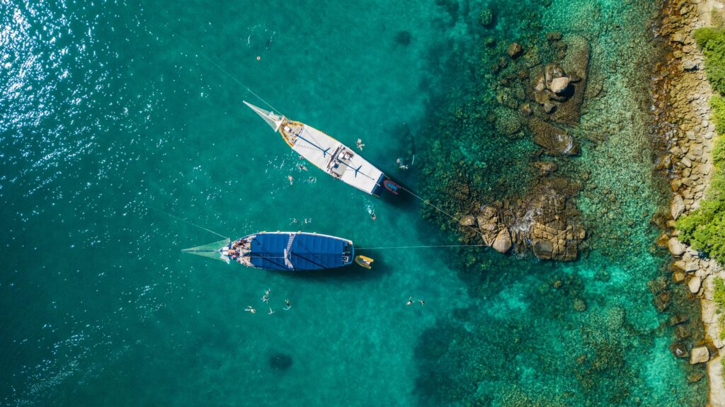 Vista aérea de dois barcos de passeio ancorados em águas verde-turquesa cristalinas de Paraty, Rio de Janeiro, com pessoas nadando ao redor e uma costa rochosa com vegetação, ilustrando a beleza e as atividades náuticas da região.