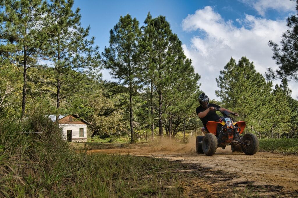 Homem pilotando um quadriciclo laranja em uma trilha de terra cercada por pinheiros e vegetação com uma pequena construção branca ao fundo e céu azul com nuvens.