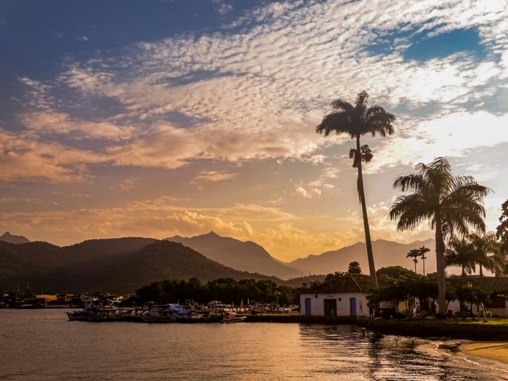 Vista panorâmica ao pôr do sol em Paraty, com montanhas verdejantes ao fundo, céu com nuvens brancas e tons alaranjados, águas calmas com barcos ancorados, palmeiras e uma construção branca com detalhes azuis na margem direita.