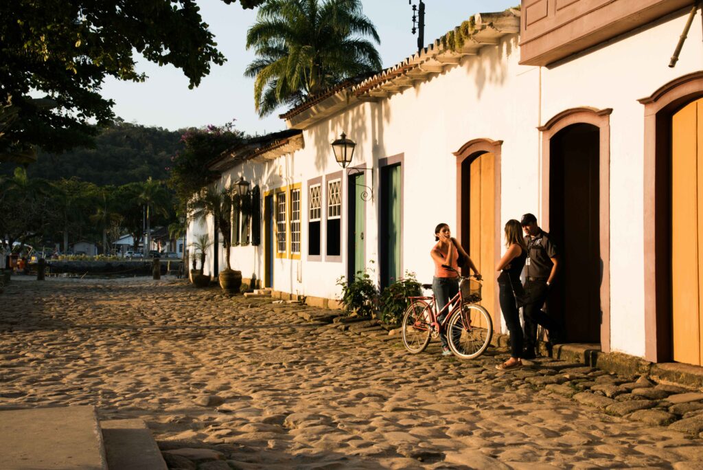 Rua de paralelepípedos no centro histórico de Paraty, Rio de Janeiro, com casarões coloniais brancos de portas e janelas coloridas sob a luz solar, uma bicicleta vermelha encostada em um muro e três pessoas conversando, com palmeiras e morros verdes ao fundo.