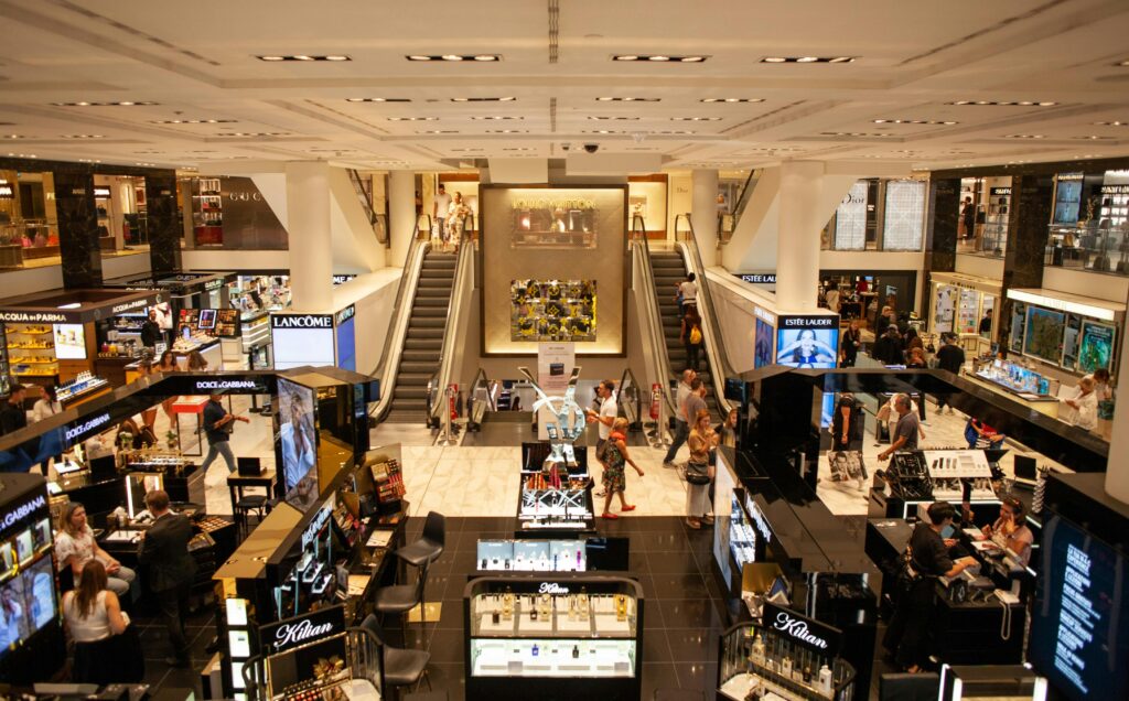 Vista panorâmica do interior de uma loja de departamento movimentada no Paraguai, com diversos stands de marcas de cosméticos como Lancôme e Dolce & Gabbana, escadas rolantes com pessoas circulando e iluminação artificial clara, representando a experiência de compra em grandes magazines paraguaias.