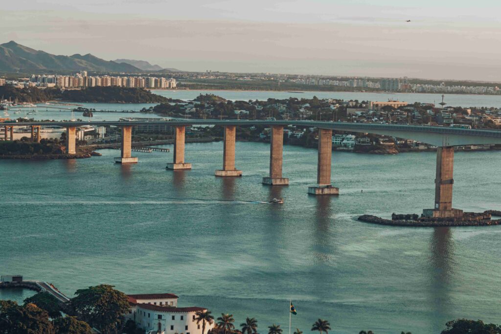 Vista aérea da Terceira Ponte em Vitória, Espírito Santo, com o mar azul-esverdeado, edifícios da cidade ao fundo e montanhas na linha do horizonte, sob um céu claro.