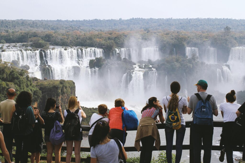 Grupo de turistas de costas observando a imensidão das Cataratas do Iguaçu no lado brasileiro, com várias quedas d'água e vegetação exuberante ao fundo, transmitindo a grandiosidade da natureza e a experiência do visitante.
