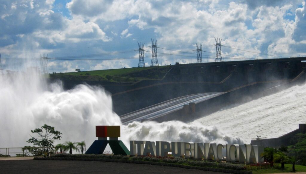 Vista panorâmica da Usina Hidrelétrica de Itaipu Binacional com o vertedouro liberando uma grande quantidade de água branca e espumante; o letreiro "ITAIPU BINACIONAL" é visível em primeiro plano, junto a uma estrutura colorida, e torres de transmissão de energia se destacam contra o céu com nuvens ao fundo.