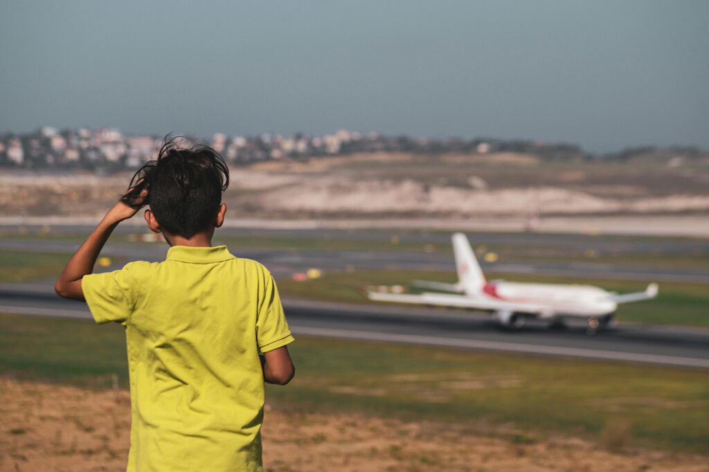 Menino de costas vestindo camiseta amarela, olhando para um avião branco com detalhes em vermelho na pista de um aeroporto, ilustrando a possibilidade de viagens aéreas para menores com a devida autorização.