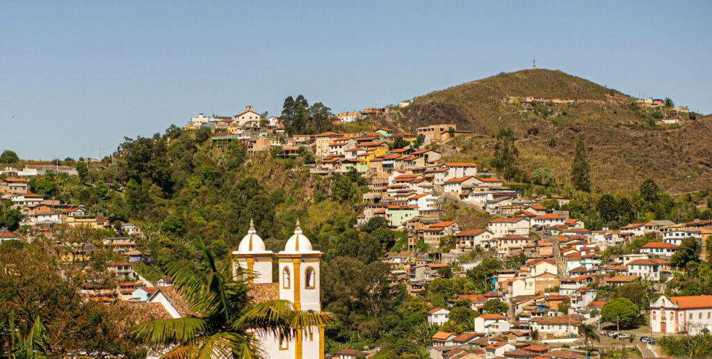 Vista panorâmica de Ouro Preto em um dia de sol, mostrando a paisagem de morros íngremes e uma série de casas coloridas. No primeiro plano, o topo de uma igreja colonial com duas torres brancas se destaca entre a vegetação e as construções, enquanto uma cruz no cume da montanha domina a paisagem.