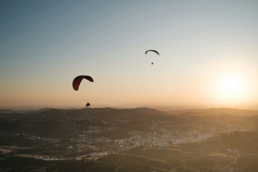 Dois parapentes coloridos voam sobre a paisagem montanhosa e a cidade de Serra Negra ao pôr do sol, com tons alaranjados no céu.