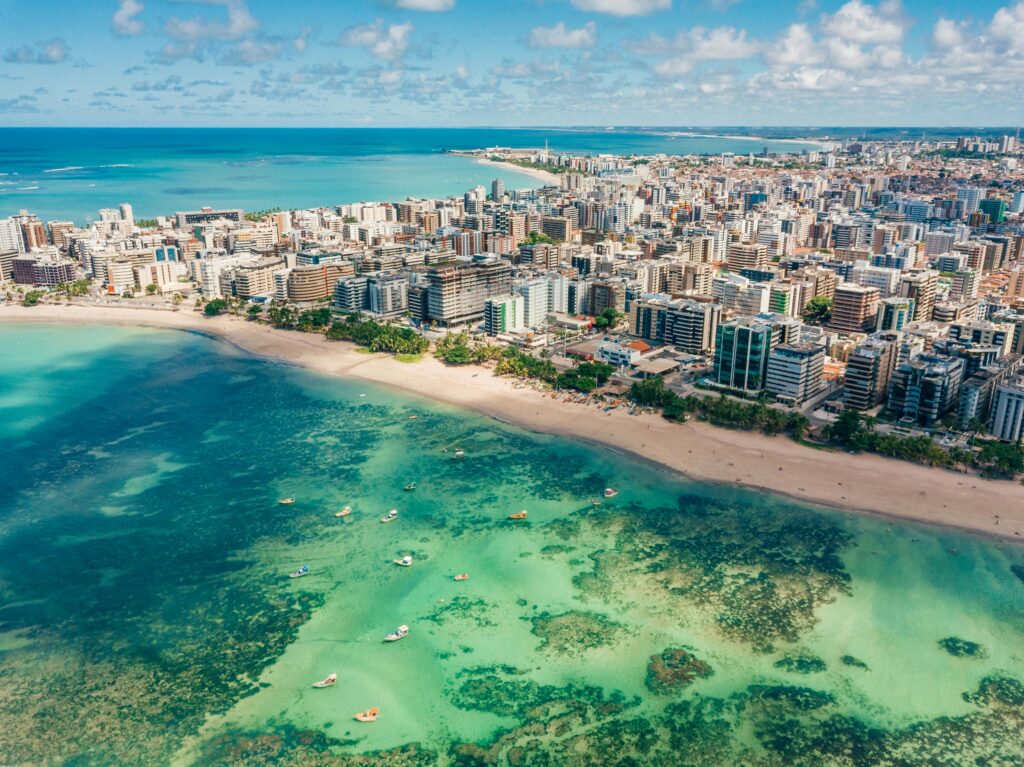 Vista aérea da orla de Maceió, em Alagoas, mostrando a praia de areia clara, o mar de águas calmas e transparentes com barcos e a densa área urbana com edifícios altos à beira-mar.