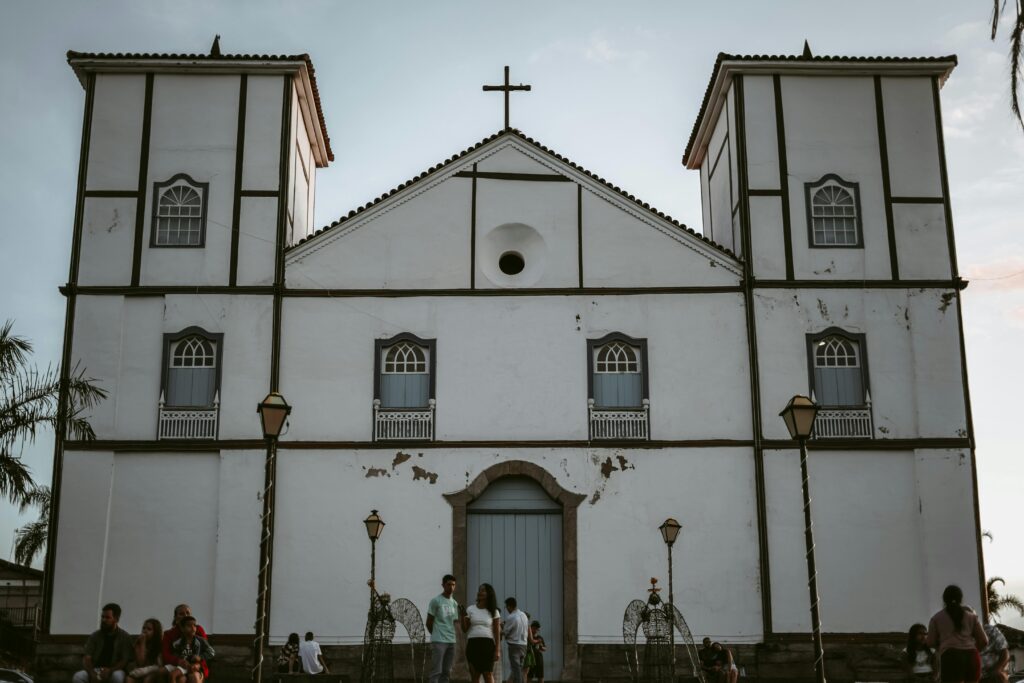 Fachada da Igreja Matriz de Nossa Senhora do Rosário, em Pirenópolis, com suas duas torres brancas, uma cruz no topo e pessoas sentadas em frente, destacando a arquitetura colonial da cidade.