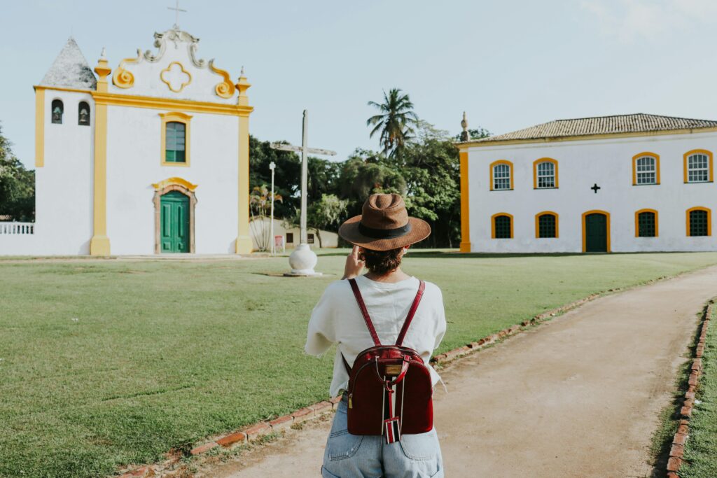 Uma turista com chapéu e mochila caminha em um caminho de terra em Porto Seguro em direção a uma igreja colonial branca e amarela e outras construções históricas.